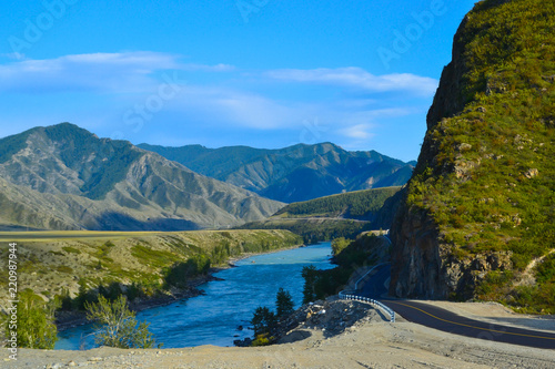 Fototapeta Naklejka Na Ścianę i Meble -  A new winding asphalt black road with a yellow dividing strip between the rocks and the Katun River in the Altai mountains under a clear blue sky with clouds and a field covered with green grass