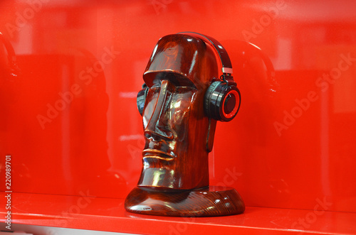 Fototapeta Naklejka Na Ścianę i Meble -  Wooden head statue in Easter Island style with headphones on head on red background on shelves shop