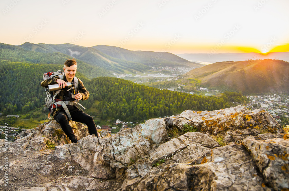 A young tourist with backpack sits on top of a hill on rocks and drinks tea from a thermos bottle against the background of the mountain and the village. A young tourist with backpack sits on top of a hill on rocks and drinks tea from a thermos bottle against the background of the mountain and the village.