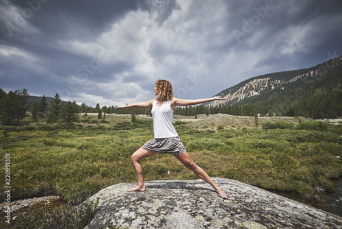 a young woman with curly hair doing yoga on top of a giant boulder in the mountains