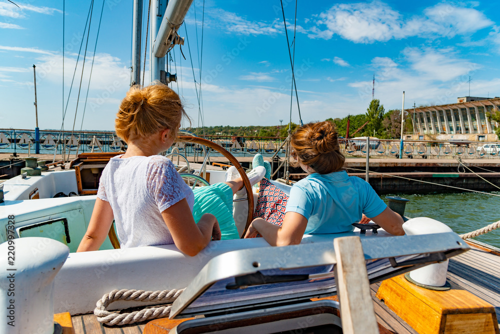 Two girls are sitting on the yacht near the helm and looking out into the sea on the deck.