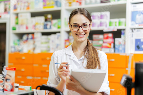A pretty slim dark-haired girl with glasses,dressed in a medical overall,writes notes in a notebook at the cash desk in a new pharmacy.