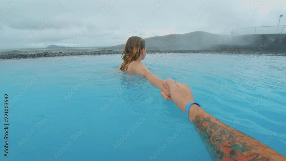 POV of follow me shot of man holding hand of woman girlfriend, walking or swimming in amazing turqouise blue water of natural hot spring or thermal swimming pool, romantic getaway in iceland