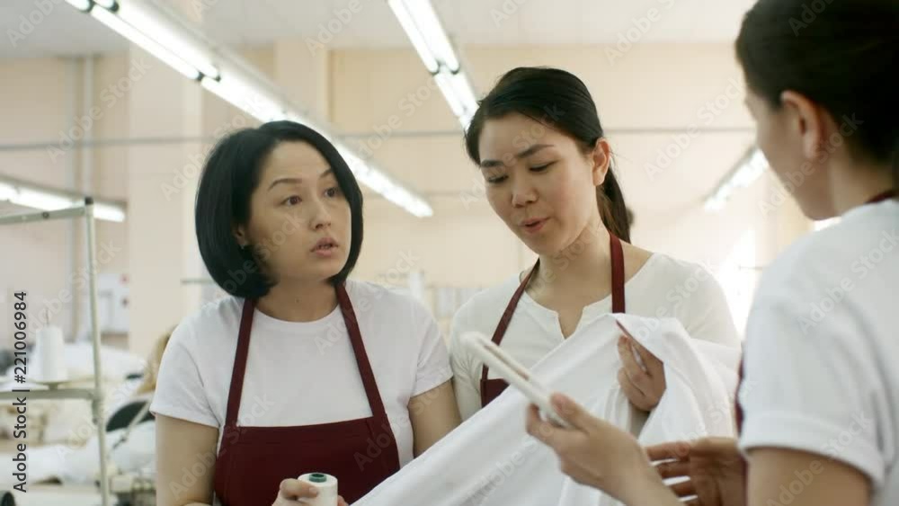 Asian seamstresses walking through sewing factory and discussing color of thread and fabric while female colleague showing something on digital tablet to them Asian seamstresses walking through sewing factory and discussing color of thread and fabric while female colleague showing something on digital tablet to them