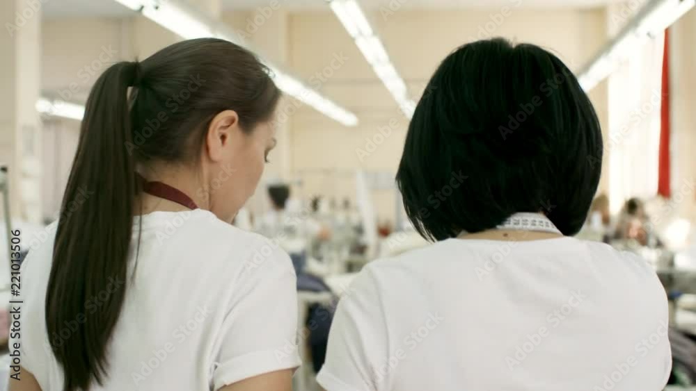 Rear view of two professional seamstresses walking through sewing factory and discussing material Rear view of two professional seamstresses walking through sewing factory and discussing material