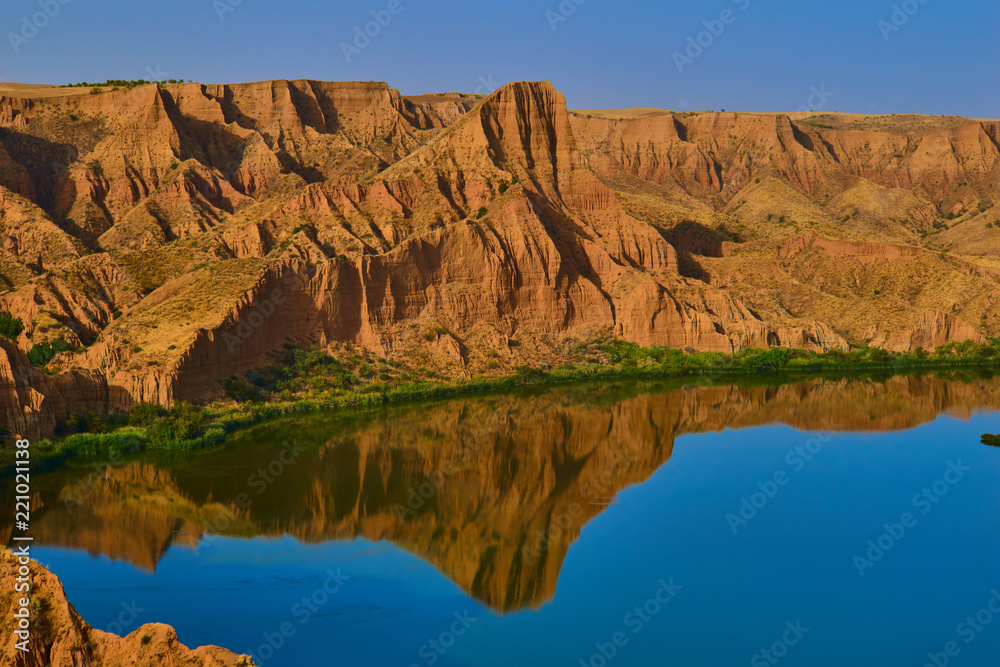 Beautiful landscape with red rocks and lake in the foreground with reflection in the water of the mountains with clear sky in the Barrancas de Burujon, Toledo, Spain
