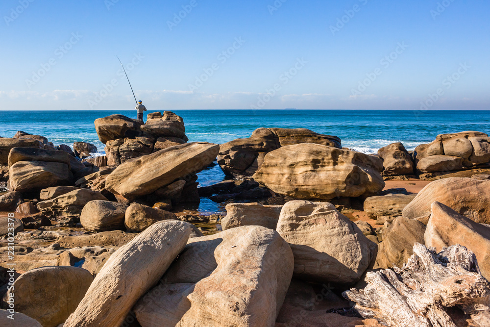 Fisherman fishing off large rock boulders on beach with with ocean towards the horizon. Fisherman fishing off large rock boulders on beach with with ocean towards the horizon.