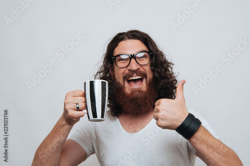 Cheerful young bearded man wearing eyeglasses showing thumbs up gesture and holding cup of coffee or tea