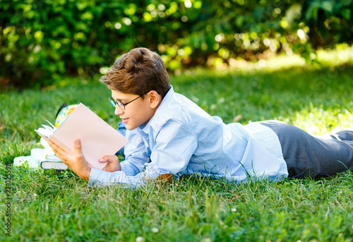 cute, young boy in round glasses and blue shirt reads book lying on the grass in the park. Education, back to school concept