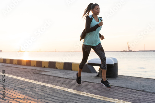 Side view of fitness woman running on a road by the sea. Sportswoman training on seaside promenade at sunset. © opolja