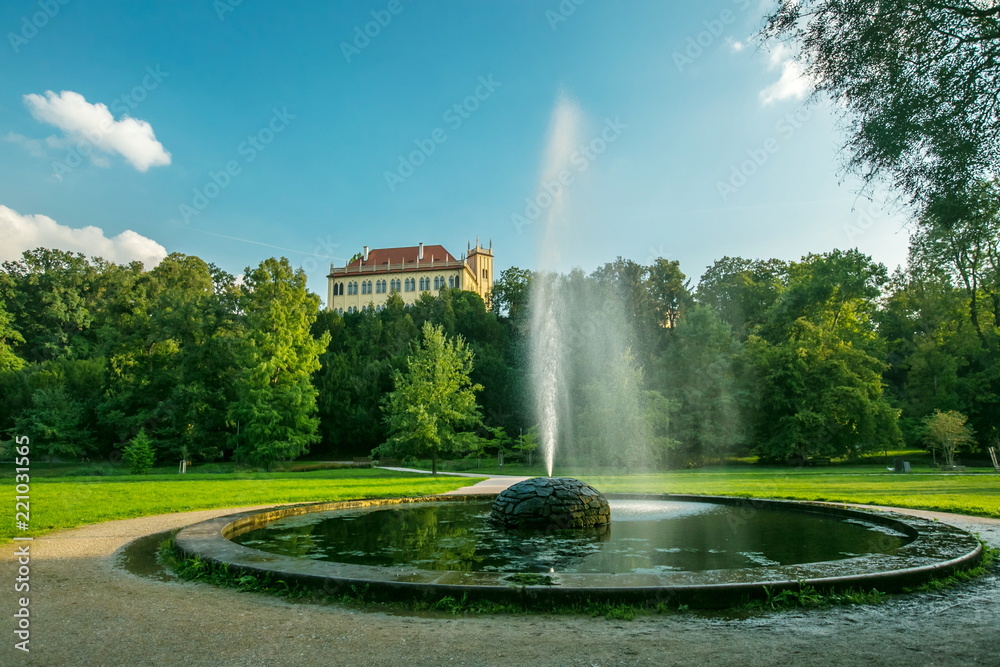 View of round water fountain with sprinkle, green lawn and trees in a famous Prague park Stromovka, yellow building of Governor’s Summer Palace from 15 century on the top of hill, summer day, blue sky