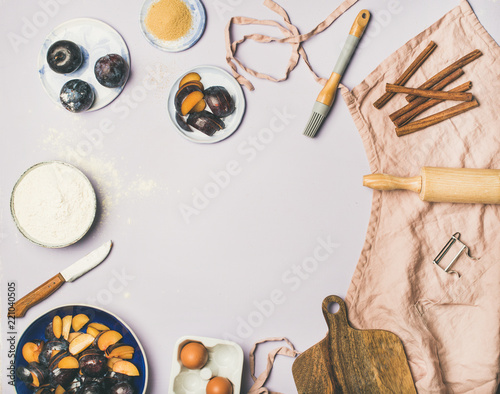 Baking ingredients and tools. Flat-lay of dusty pink linen apron, kitchen utensils, flour, sugar, fresh plum, egg and spices over pastel lilac background, top view, copy space