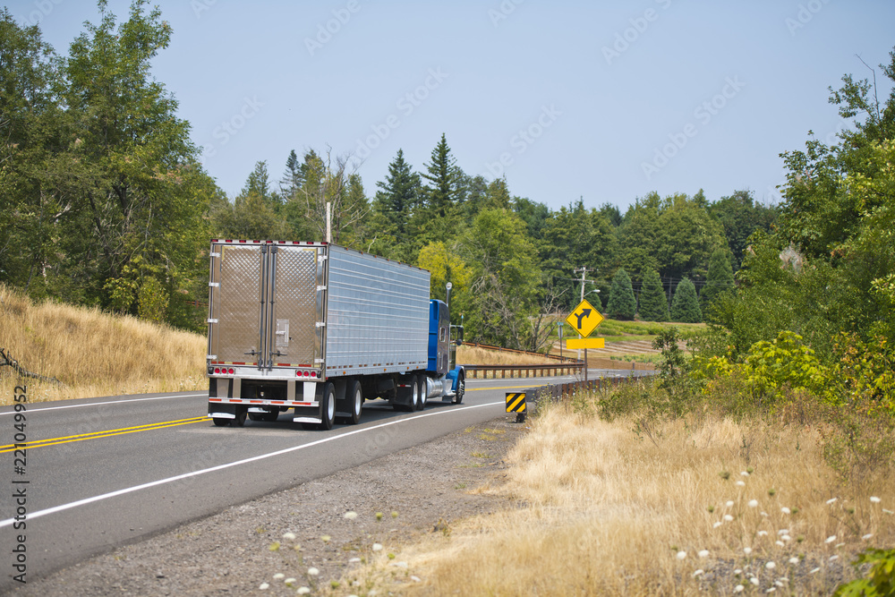 Blue big rig semi truck with refer semi trailer running on winding green highway for delivery
