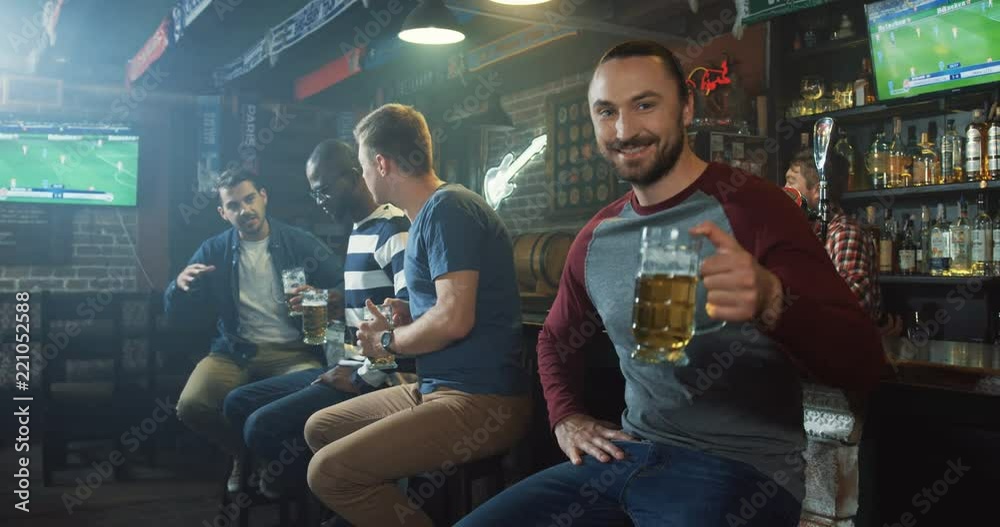 Portrait shot of the young Caucasian man watching sport match at the bar, then doing cheers with beer to the camera and smiling.