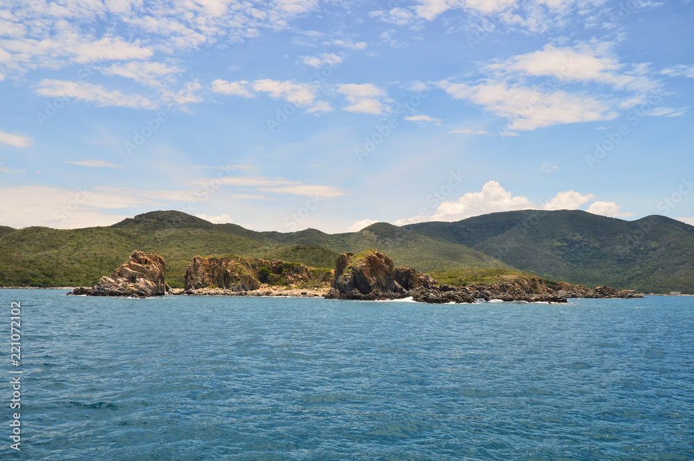 island covered with tropical vegetation in the South China Sea, coastline, hills, against a blue sky covered with clouds