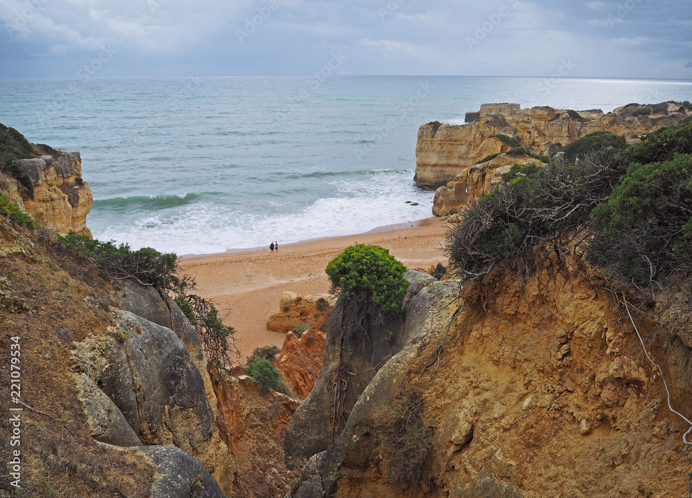 beautiful sandstone cliffs with view on two people standing on sand beach with sea waves and rock formation and blue sky background in Algarve
