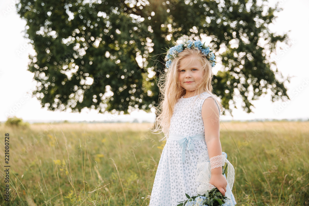 Little girl in sky blue dress with wreath and bouquet stand in field in front of big tree Little girl in sky blue dress with wreath and bouquet stand in field in front of big tree