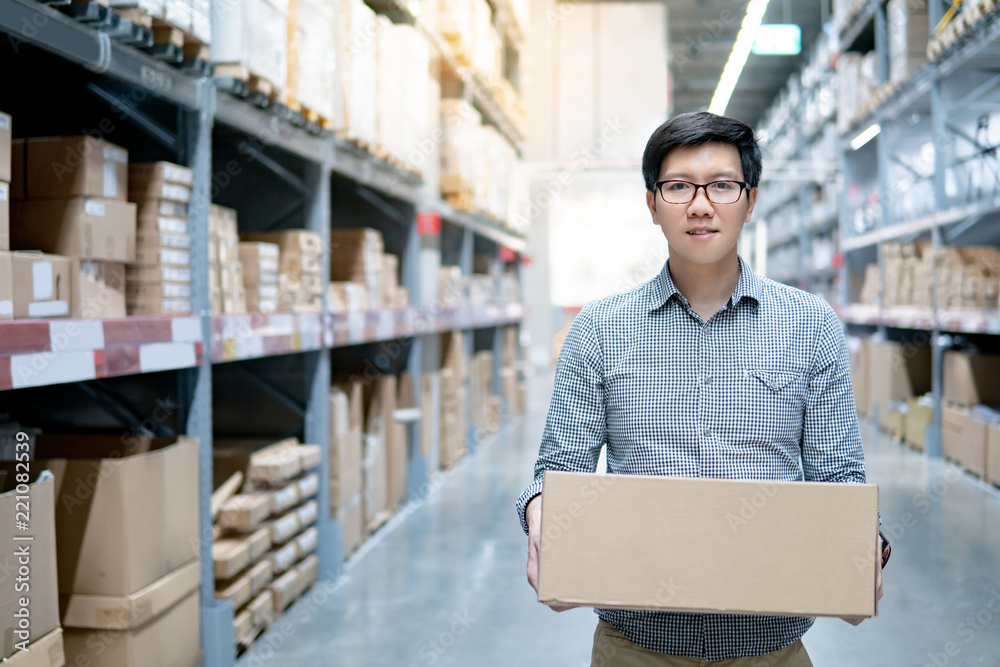 Young Asian man carrying cardboard box between row of shelves in warehouse. Shopping warehousing or working pick and packing concepts Young Asian man carrying cardboard box between row of shelves in warehouse. Shopping warehousing or working pick and packing concepts