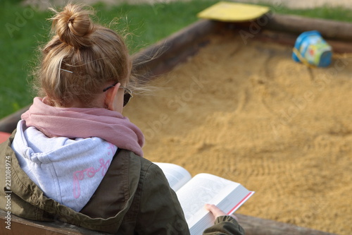 Nastolatka (dziewczynka, młoda kobieta) siedzi na skraju piaskownicy i czyta grubą książkę, blond włosy ma upięte w kok, różowy szalik, zieloną kurtkę, okulary słoneczne, widok z boku