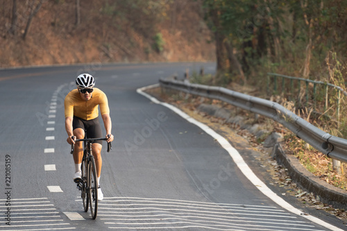 Focus on Asian man wearing a yellow cycling jersey, who's riding a road bike up high on hill in the morning. Under morning sunshine with determination on his face. © Mongkol