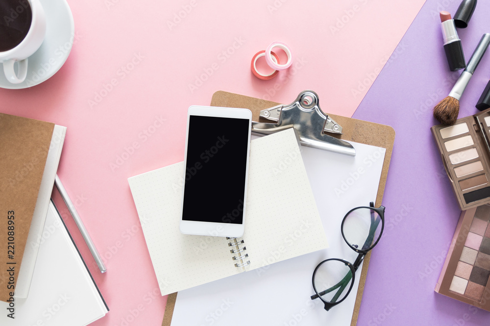 Top view over the beauty working desk with opened notebooks, cup of coffee, pen, make up brush, lipstick, eye shadows, paper, glasses and phone all over the desk. Copy space for text.