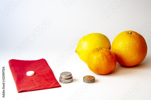 Pill on a red serviete, lemon, orange, mandarin orange and organized money coins for comparison isolated over white. Health and medical concept.