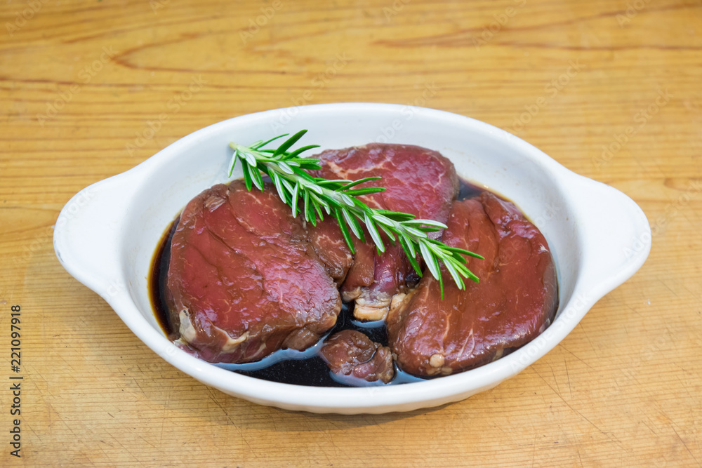 Meat marinating: Beef steak eye fillet in white dish of soy sauce marinade and rosemary herb sprig on wooden background with copy space