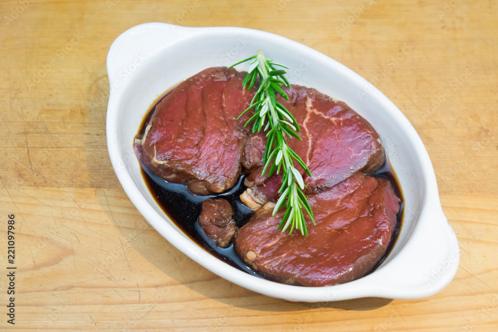 Meat marinating: Beef steak eye fillet in white dish of soy sauce marinade and rosemary herb sprig on wooden background - diagonal composition