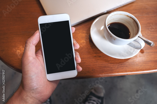 Top view mockup image of hand holding white mobile phone with blank black desktop screen with coffee cup and laptop on wooden table in cafe