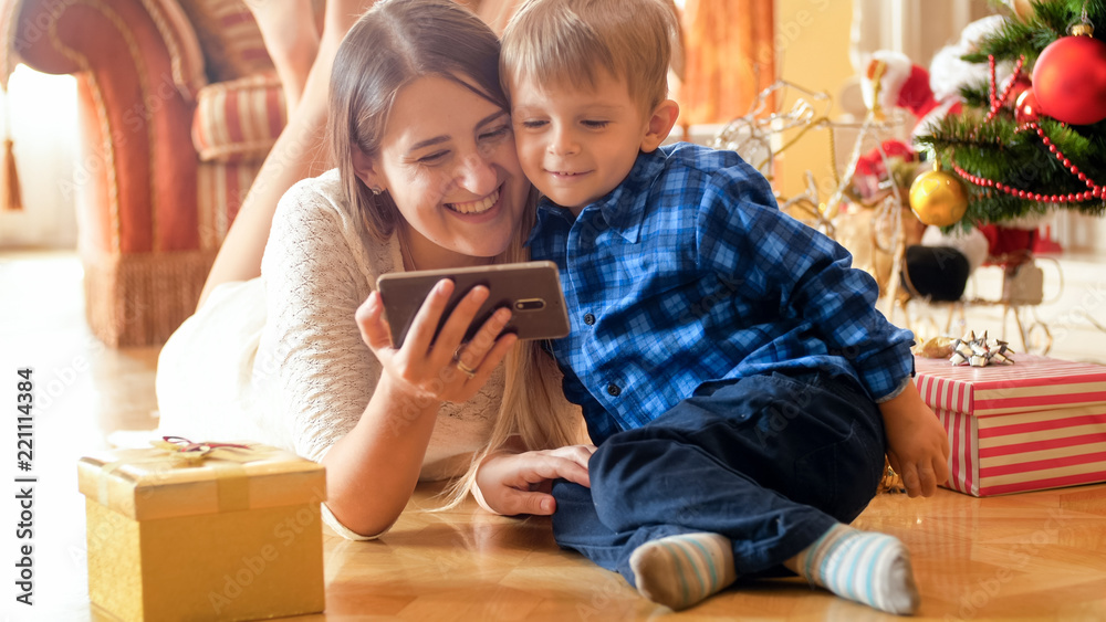 Portrait of cheerful smiling mother with baby lying on floor at living room and watching video on smart phone Portrait of cheerful smiling mother with baby lying on floor at living room and watching video on smart phone