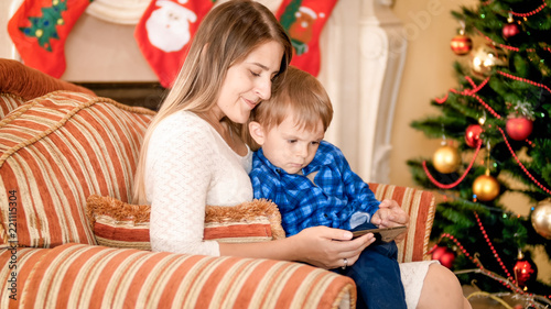 Beautiful young mother sitting in armchair with her little boy next to Christmas tree and watching video on smartphone
