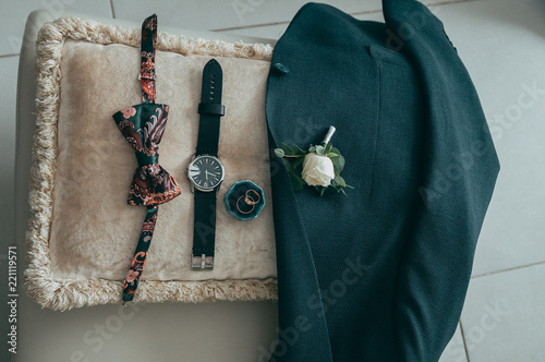 Groom's accessories: a watch, a green jacket, a buttonhole, a butterfly and rings on a tinted beige cushion.