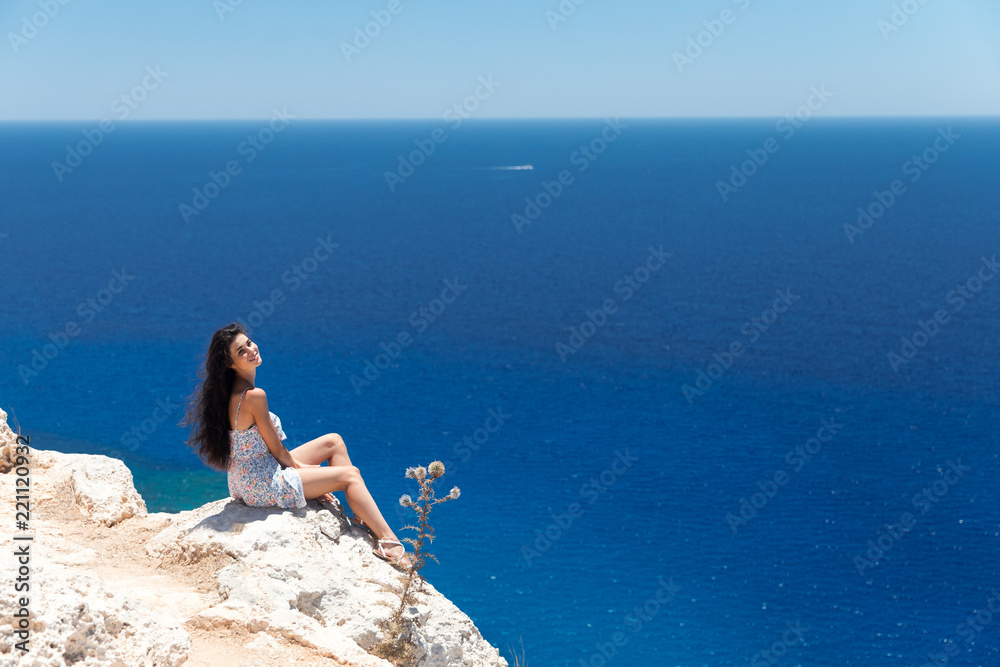 Brunette in a dress sits on the edge of the cliff overlooking the sea