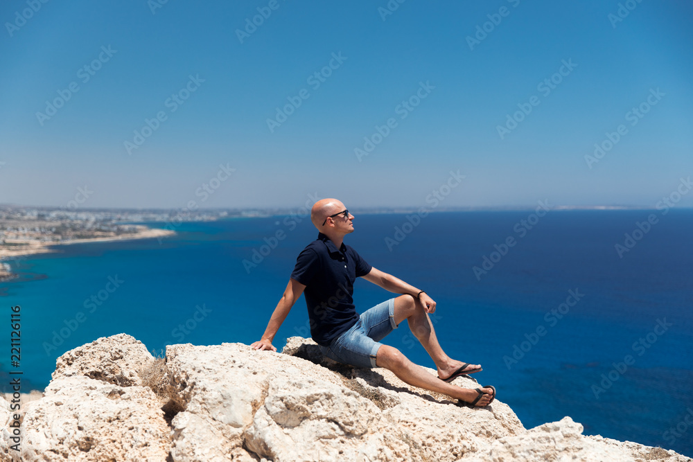 Bald man in a T-shirt and shorts sitting alone on top of a mountain overlooking the sea Bald man in a T-shirt and shorts sitting alone on top of a mountain overlooking the sea