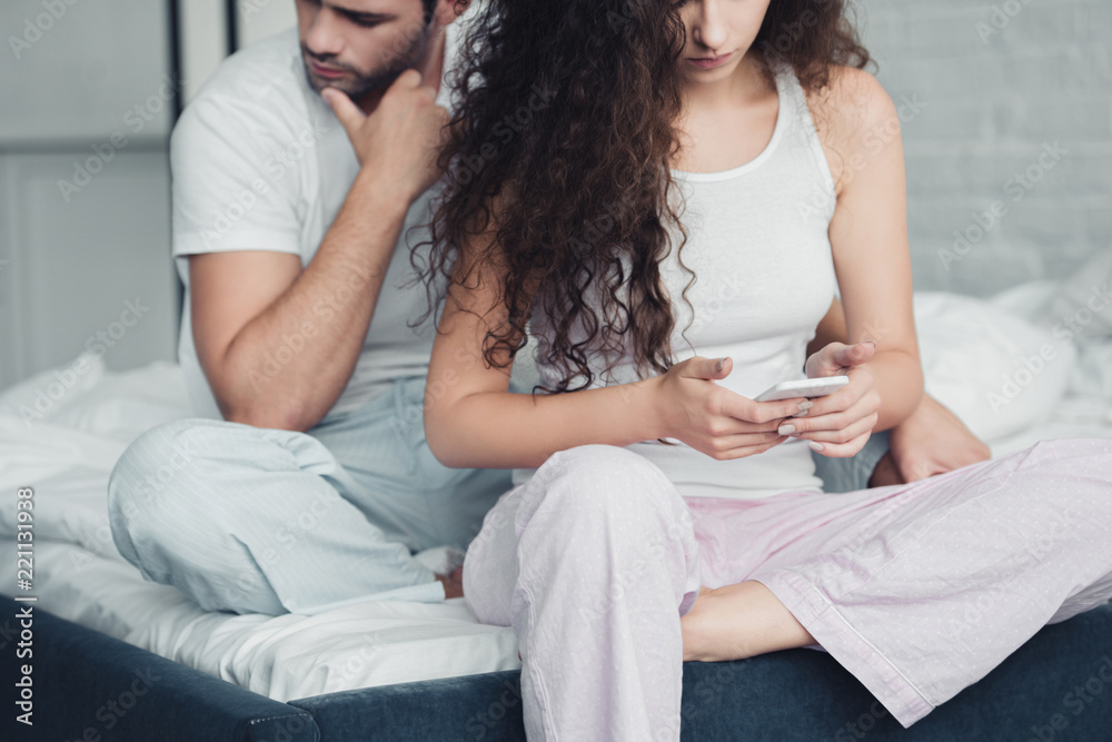cropped shot of young woman using smartphone while upset man sitting behind on bed, relationship difficulties concept