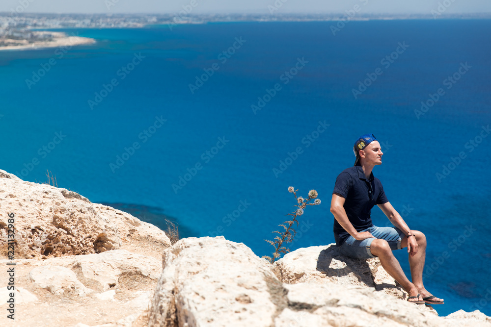 Bald man in a T-shirt and shorts sitting alone on top of a mountain overlooking the sea