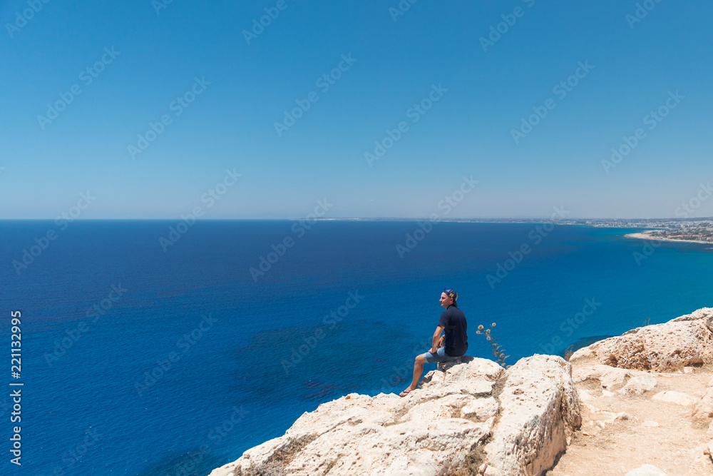 Bald man in a T-shirt and shorts sitting alone on top of a mountain overlooking the sea Bald man in a T-shirt and shorts sitting alone on top of a mountain overlooking the sea