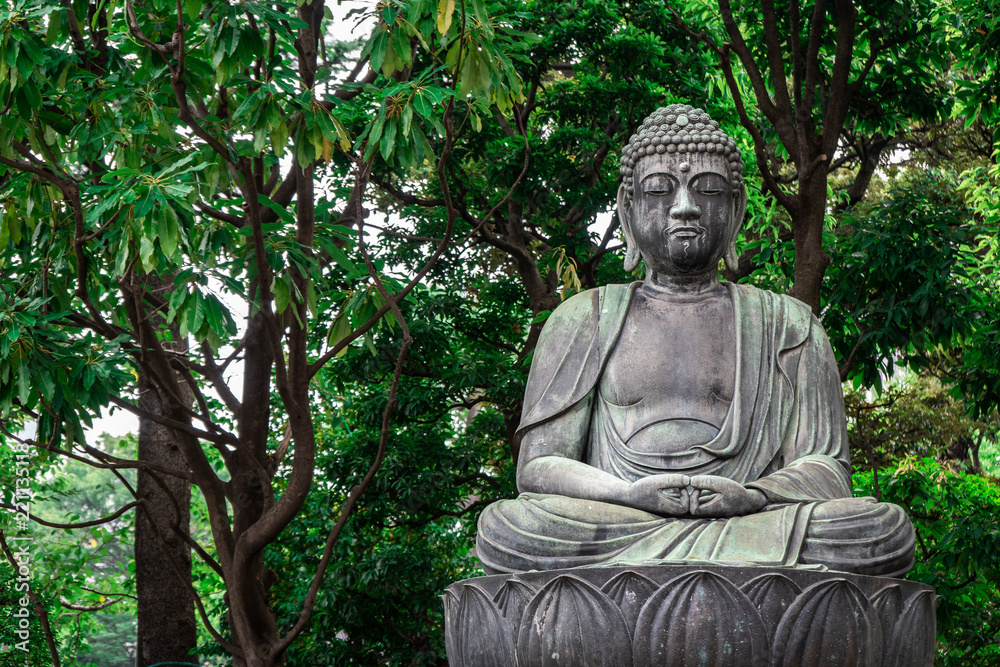 Stone statue of sitting Buddha on a lotus leaf with green trees in the background, Tokyo, Japan Stone statue of sitting Buddha on a lotus leaf with green trees in the background, Tokyo, Japan