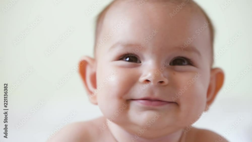 A cute little baby is looking into the camera and is happy on a white bed sheet smiles close up portrait A cute little baby is looking into the camera and is happy on a white bed sheet smiles close up portrait