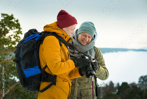 Two mature men with exploring Finland in winter. Travellers hiking and Taking pictures with camera on the top of rock. Beautiful view of northern landscape with frozen Baltic Sea and snowy islands. © Suzi Media 