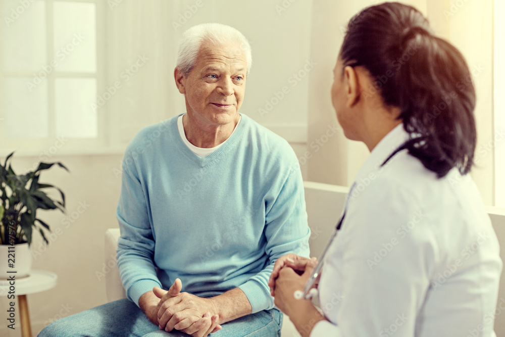 Preventive consultation. Selective focus on a cheerful senior gentleman sitting on a sofa and listening to a female medical worker during a checkup. Preventive consultation. Selective focus on a cheerful senior gentleman sitting on a sofa and listening to a female medical worker during a checkup.