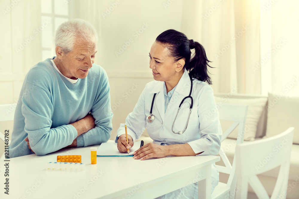 Fototapeta premium Planning the course of treatment. Positive minded female nurse and her senior patient sitting at a table and discussing the treatment during a consultation.