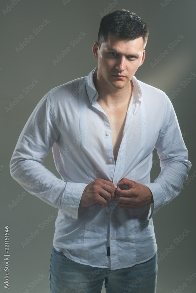 Man unbutton fashionable shirt. Young macho undress white shirt. Guy with serious face. Sexy model with elegant casual style. Fashion striptease and style concept Man unbutton fashionable shirt. Young macho undress white shirt. Guy with serious face. Sexy model with elegant casual style. Fashion striptease and style concept