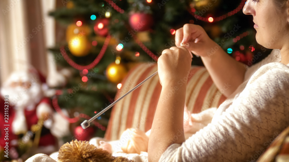 Closeup image of young woman sitting by the CHristrmas tree and knitting woolen scarf