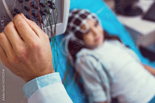 Getting ready. Scaled up look on a hand of a laboratory worker setting up an electroencephalography machine before analyzing brain of a little girl lying on an examination couch in the background.