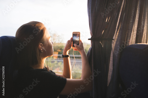 Tourist woman sits in a bus near the window and photographs landscapes at sunset on a smartphone. Tourist picks up landscapes from the bus window on a smartphone. Travel by bus.