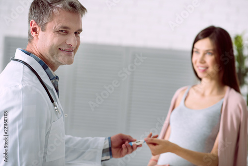 Temperature check. Delighted nice positive doctor holding a thermometer and smiling while giving it to his patient