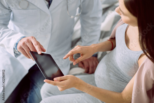 Useful device. Modern tablet being in hands of a nice positive pregnant woman while sitting together with her doctor