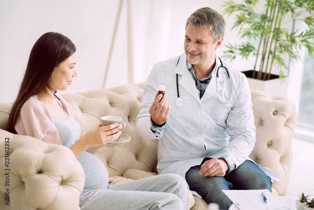 Fototapeta premium Take this. Happy smart cheerful doctor holding a bottle with medicine and smiling while giving it to his patient