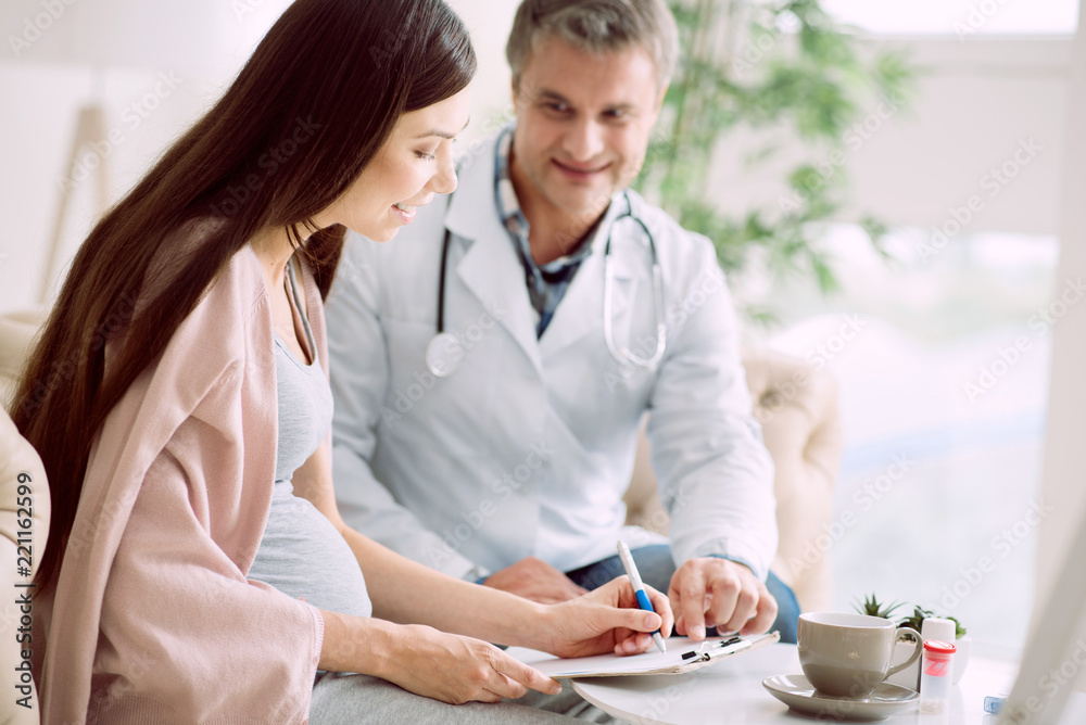 Medical agreement. Joyful positive pregnant woman smiling and signing the document while sitting together with her doctor Medical agreement. Joyful positive pregnant woman smiling and signing the document while sitting together with her doctor